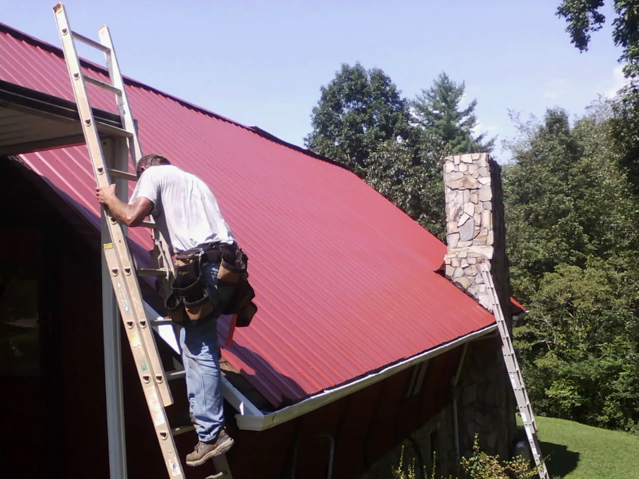 Roofer on a ladder beside a red metal roof and stone chimney — Soffit & Fascia Repair by Roofing Brothers in Hasbrouck Heights